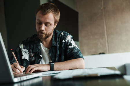 Photo of serious handsome man working with laptop and writing down notes while sitting at table...