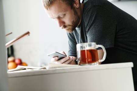 Photo of serious redhead man with beard using mobile phone while drinking tea in kitchen at home