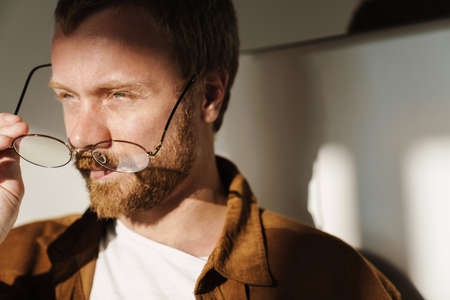 Photo closeup of thinking young man holding eyeglasses and looking aside indoors