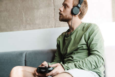 Photo of focused man wearing headphones playing video game with joystick while sitting on sofa at homeの写真素材