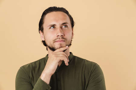 Image of young serious man thinking and looking aside at copyspace isolated over beige background