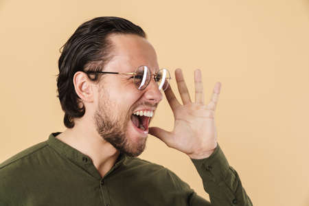 Image of young annoyed man in eyeglasses screaming and looking aside isolated over beige backgroundの写真素材