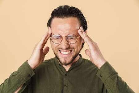 Image of young unhappy man with headache in eyeglasses rubbing his temples isolated over beige...