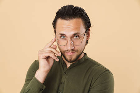 Image of young confident man wearing eyeglasses looking at camera isolated over beige background