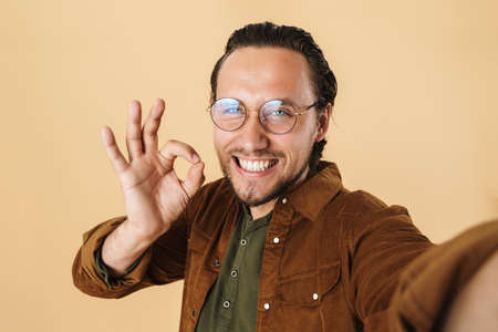 Image of happy man in eyeglasses gesturing ok sign while taking selfie photo isolated over beige...