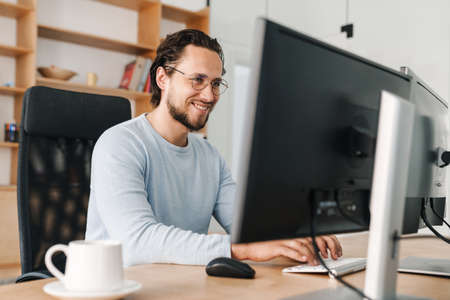 Image of smiling unshaven programmer man wearing eyeglasses working with computer in officeの写真素材