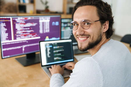 Image of cheerful caucasian programmer man wearing eyeglasses working with computers in office