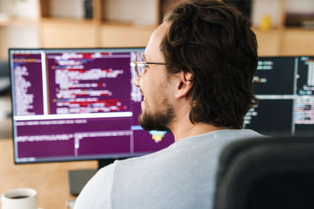 Image of unshaven pleased programmer man wearing eyeglasses working with computers in office