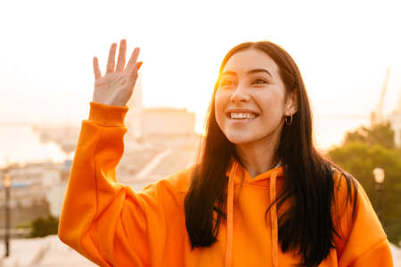 Photo of joyful beautiful asian woman smiling and waving hand while walking outdoorsの写真素材