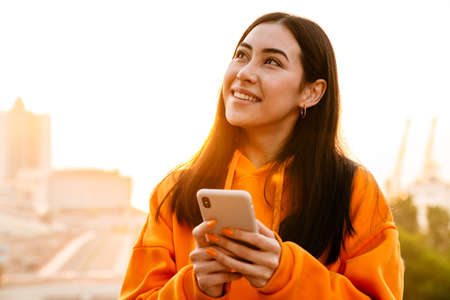 Photo of joyful attractive asian woman smiling and using mobile phone while walkingoutdoors