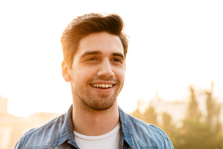 Photo of joyful unshaven man in denim shirt smiling and looking aside while walking outdoorsの写真素材
