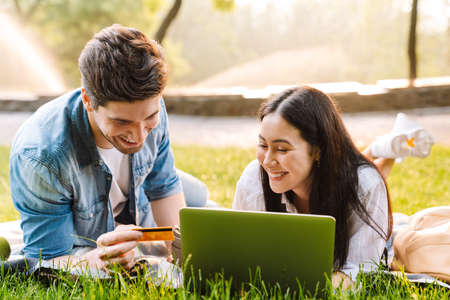 Image of joyful multicultural couple using laptop and holding credit card while lying on grass in...