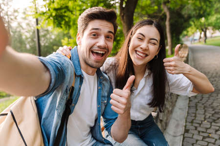 Image of joyful multicultural couple taking selfie photo and showing thumbs while sitting on bench in parkの写真素材