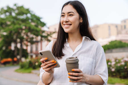 Photo of happy asian woman using cellphone and drinking coffee takeaway while walking on boulevardの写真素材