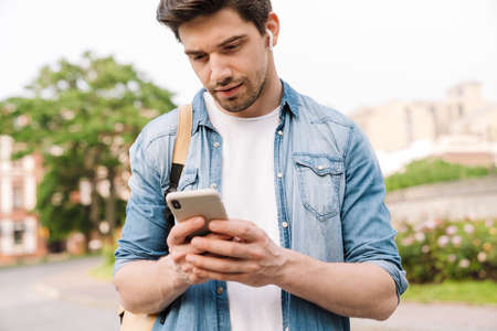 Photo of focused man with wireless earphone using cellphone while walking with backpack on boulevard