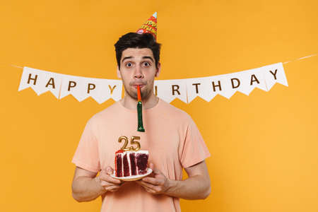 Photo of surprised man holding birthday cake and blowing in whistle isolated over yellow backgroundの写真素材