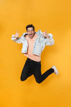 Photo of delighted handsome man jumping and showing thumb up isolated over yellow background