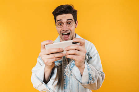 Photo of excited young man playing online game on mobile phone isolated over yellow backgroundの写真素材