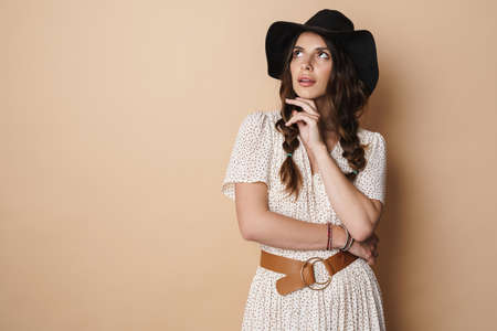 Photo of thinking beautiful woman in hat posing and looking upward isolated over beige background
