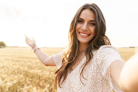 Photo of beautiful happy woman in stylish dress smiling while taking selfie photo on wheat field