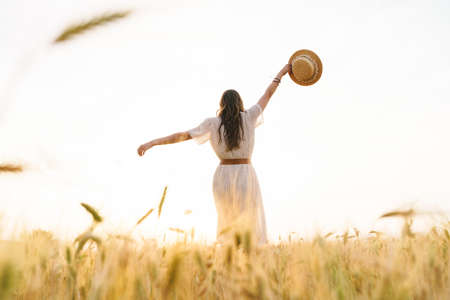 Photo from back of cute brunette woman with straw hat dancing on wheat field at summer dayの写真素材