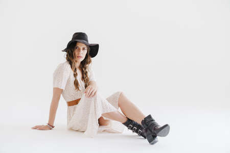 Photo of brunette seductive woman in hat posing and sitting on floor isolated over white background