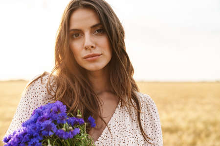 Photo of beautiful cute woman in stylish dress posing while walking with flowers on wheat fieldの写真素材