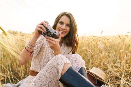 Photo of charming joyful woman in stylish dress using retro camera while sitting on wheat field