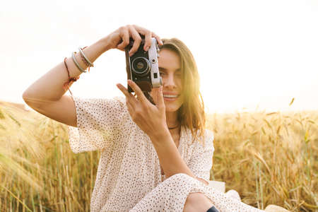 Photo of charming joyful woman in stylish dress using retro camera while sitting on wheat field