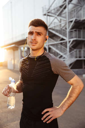 Image of young athletic sportsman in earphones drinking water while working out at urban area...