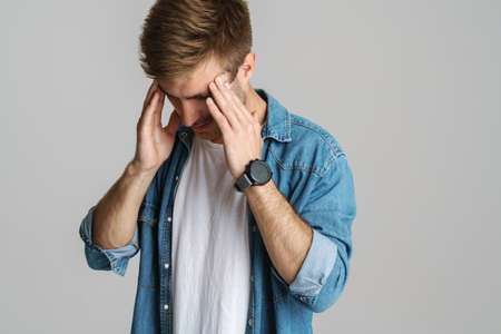 Portrait of unhappy young man with headache rubbing his temples isolated over grey backgroundの写真素材