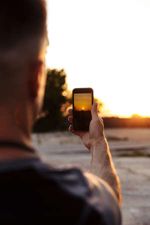 Image from back of sportsman taking photo on mobile phone while working out on sunrise outdoors