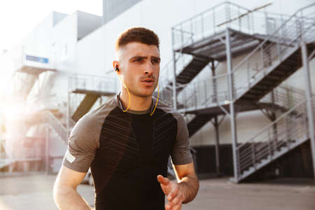 Image of focused caucasian sportsman in earphones running while working out at urban area outdoors