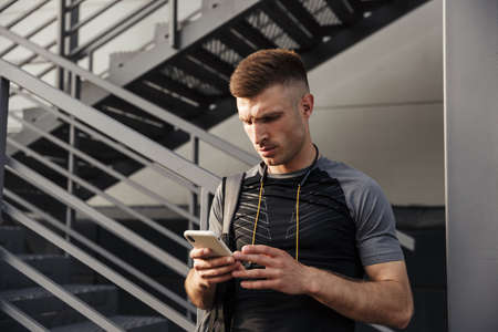 Image of young athletic sportsman with earphones using cellphone while working out near urban stair...