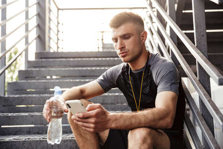 Image of unshaven caucasian sportsman drinking water and using cellphone while sitting on urban...