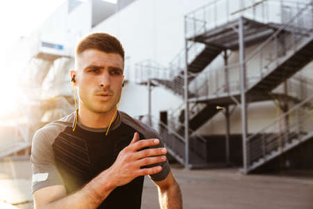 Image of focused caucasian sportsman in earphones running while working out at urban area outdoors