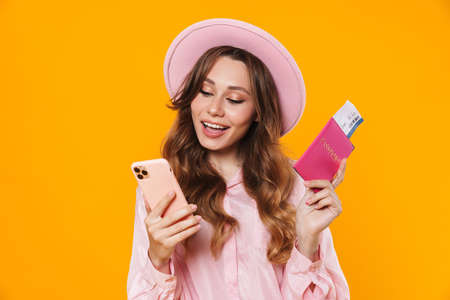 Image of smiling woman in hat holding passport with ticket and mobile phone isolated over yellow backgroundの写真素材