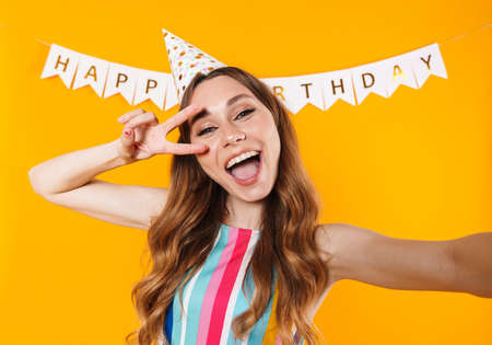 Image of joyful cute woman taking selfie photo and gesturing peace sign isolated over yellow backgroundの写真素材