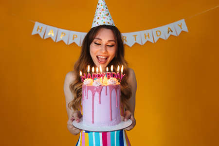 Image of delighted young woman in party cone showing birthday torte with candles isolated over yellow backgroundの写真素材