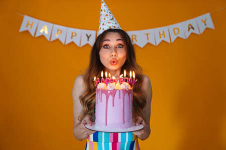 Image of happy young woman in party cone blowing out candles on birthday torte isolated over yellow backgroundの写真素材