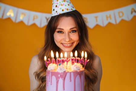 Image of delighted young woman in party cone showing birthday torte with candles isolated over yellow backgroundの写真素材