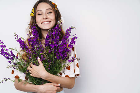 Photo of cheerful woman with fake butterflies and flowers smiling at camera isolated over white wallの写真素材