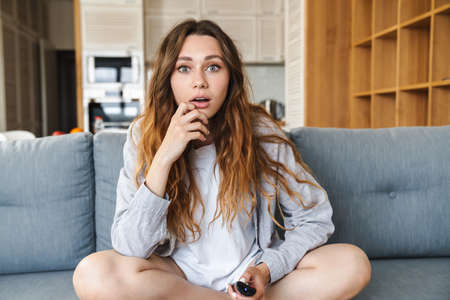 Shocked young woman relaxing on a couch at home, holding tv remote controlの写真素材