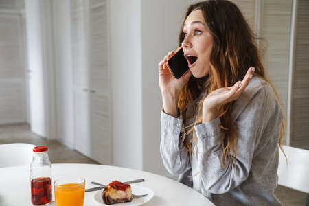 Photo of excited beautiful woman talking on cellphone while having breakfast in white kitchenの写真素材
