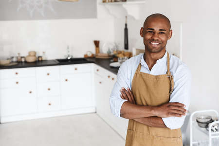 Photo of cheerful african american man in apron smiling while posing with arms crossed at kitchen