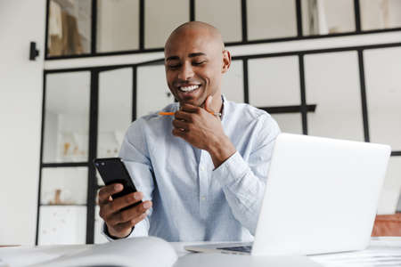 Photo of laughing african american man using mobile phone while working with laptop at table in...