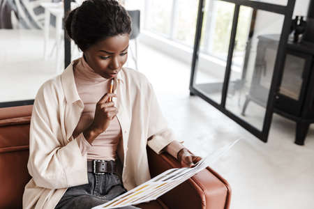 Photo of pretty concentrated young african woman sitting on sofa indoors at home while holding catalogue with colorsの写真素材