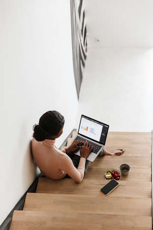 Top view of an attractive young african woman sitting on a staircase at home, working on laptop...
