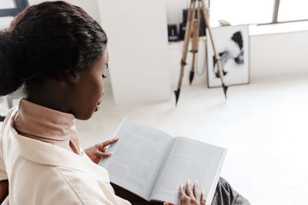 Side view photo of young african woman sitting on sofa indoors at home while reading magazine or bookの写真素材