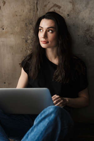 Photo of beautiful young brunette woman in jeans sitting on floor and using laptop isolated over...